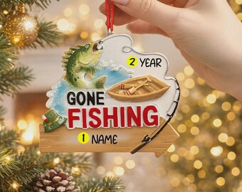 Könnte beinhalten: Weihnachtsschmuck mit Angelmotiv. Der Schmuck zeigt einen grünen Fisch, ein kleines Boot und die Worte "GONE FISHING" in Rot. Der Schmuck wird an einer roten Schnur gehalten.
