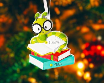 Op de afbeelding: Een groen en geel rupsornament met een bril, bovenop een stapel boeken. Het bovenste boek is open en toont de naam "Laney". Op het onderste boek staat het woord "YEAR". Een feestelijke vakantiedecoratie.