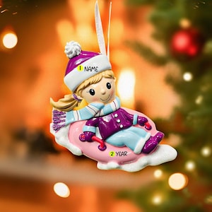 May include: A Christmas ornament featuring a girl in winter attire, including a purple hat and coat, a blue scarf, and boots. The ornament is on a pink sled, with the text "NAME" and "2 YEAR" visible. The background is blurred with Christmas decorations.