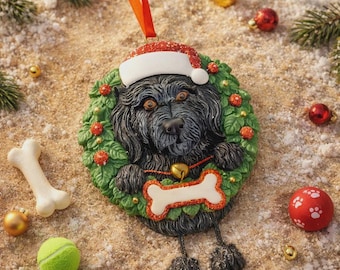 May include: A festive Christmas ornament featuring a black dog wearing a Santa hat and a wreath. The dog has a bone-shaped tag and is surrounded by Christmas decorations, including a bone, a tennis ball, and red and gold baubles.