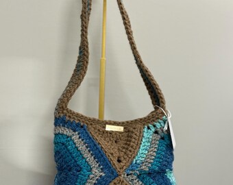 May include: A handmade crochet shoulder bag in shades of brown, blue, and grey. The bag features a geometric pattern on the front and a long strap. A small gold tag is attached to the bag.