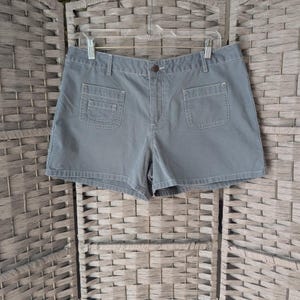 May include: Gray shorts with two front pockets and a button closure. The shorts are hanging on a metal hanger against a woven background. The shorts have a classic, casual style.