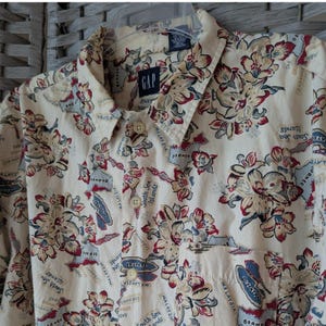 May include: Cream-colored button-down shirt with a floral and map print. The shirt features a collar, a chest pocket, and the "GAP" logo. The print includes red, blue, and beige floral designs and map outlines.
