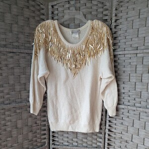 May include: Cream-colored sweater with gold sequin fringe detail across the shoulders and neckline. The long-sleeved sweater has a scoop neck and a ribbed hem and cuffs. The label reads "Maurada".
