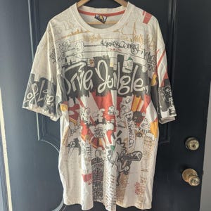 May include: White t-shirt with a colorful graphic design featuring the words "Drive Jungle" in large letters. The design includes cartoon figures, cityscapes, and graffiti-style text in black, red, and yellow.