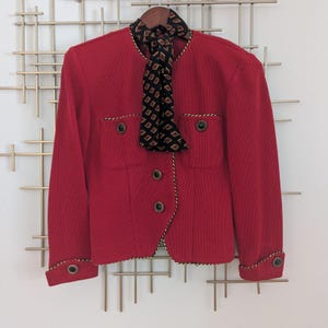 May include: A red blazer with a black and brown patterned scarf. The blazer has a round collar, two front pockets, and gold trim. The blazer has three black buttons down the front and one on each cuff.