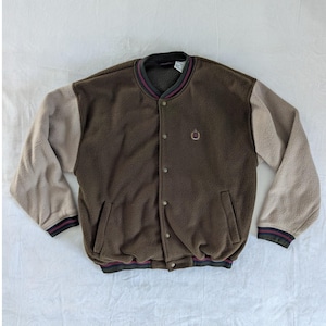 May include: Brown fleece jacket with tan sleeves. The jacket has a button-up front, two pockets, and a small embroidered emblem. The collar and cuffs have striped detailing in black, burgundy, and green.