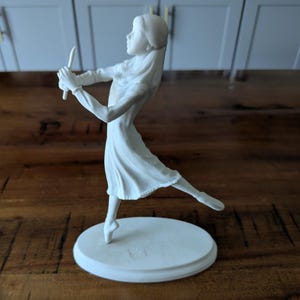 May include: A matte white porcelain figurine of a person in a dress, captured in a dynamic pose with one leg extended backward and arms raised while holding a slender, tapered object. The figure stands on an oval base with a smooth finish.