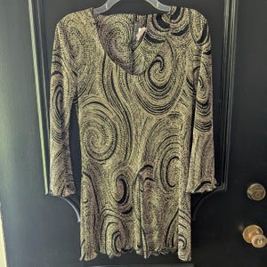 May include: A long-sleeved tunic top featuring a black and metallic gold swirling spiral pattern. The garment has a draped cowl neckline and a slightly flared hemline, with a textured fabric finish throughout the entire piece.