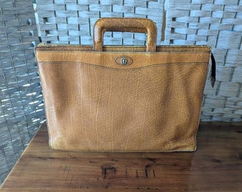 May include: A tan leather briefcase with a textured surface, featuring a rectangular top handle and a central silver-toned metal emblem on the front. The bag has a zipper closure along the top edge and a structured, rectangular silhouette.