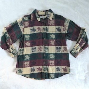 May include: A long-sleeve button-down shirt featuring a checkered pattern in shades of dark green, burgundy, and cream. The fabric is decorated with a repeating print of leaves and fruit. A single chest pocket is visible on the left side.