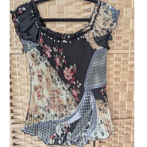 May include: A black, beige, and floral patterned top with a pleated design. The top features a square neckline with ruffled trim and sequin accents. The sleeves are short and ruffled, and the fabric has a mix of floral and geometric patterns.