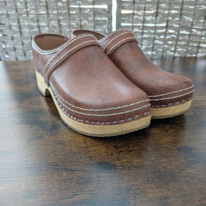 May include: A pair of brown leather clogs featuring a distressed finish, a rounded toe, and a thick wooden sole. The upper part of the shoe is accented with double rows of light-colored stitching along the base and across the top strap.
