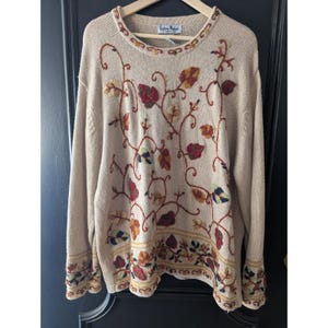 May include: Beige sweater with embroidered autumn leaves in red, brown, and green. The long-sleeved sweater has a crew neck and a decorative border at the hem and cuffs. The brand name "Extra Point" is visible on the neckline.