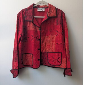 May include: A red button-down jacket featuring a patchwork-style design with various dark red and black floral and geometric patterns. The jacket includes a collared neckline, long sleeves with cuffs, and two front pockets, all outlined with black piping.