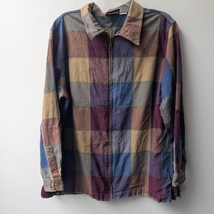 May include: A long-sleeve zip-up shirt featuring a patchwork grid pattern in muted tones of blue, purple, tan, and gray. The garment has a standard collar, a full-length front zipper, and buttoned cuffs, with visible stitching between each square.