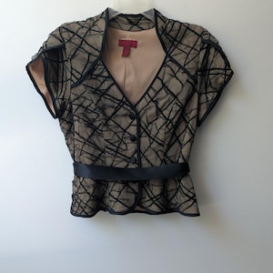May include: A short-sleeved jacket featuring a tan base with a black, crisscross, textured overlay. The jacket has a V-neckline, three black buttons down the front, and a solid black waistband. A red label is visible at the inner neckline.