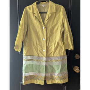 May include: A long, yellow coat with three white buttons and a collared neckline. The bottom of the coat features horizontal stripes of green, white, and yellow with floral embroidery. The coat is hanging on a black hanger.