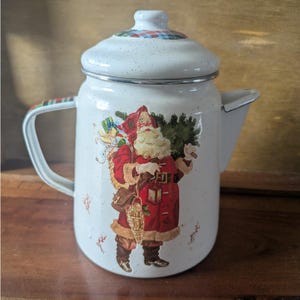 May include: White enamelware coffee pot with a Santa Claus graphic. The pot features a lid with a plaid pattern and a handle with a matching design. The Santa Claus illustration shows him carrying gifts and a Christmas tree. The pot has a spout for pouring.