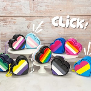 May include: A collection of heart-shaped accessories in various colors and patterns, each representing different LGBTQ+ pride flags. The accessories have a black border and a small chain. The word "Click!" is written in white.