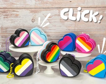 May include: A collection of heart-shaped accessories in various colors and patterns, each representing different LGBTQ+ pride flags. The accessories have a black border and a small chain. The word "Click!" is written in white.