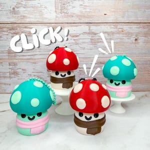 May include: Four whimsical mushroom figurines with smiling faces, each with a different colored cap: red, teal, and green. The mushrooms have white polka dots, brown scarves, and are displayed on white pedestals. The word "CLICK!" is written above.