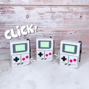 May include: Three white handheld game console keyrings with black buttons and pink accents. Each keyring has a small screen with a green glow. The word "CLICK!" is written in white above the keyrings.