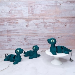 May include: Three teal-colored articulated sea creature figurines with green eyes. One figurine is on a white pedestal, while the other two are lying flat. Each has a segmented body and a black beaded chain.