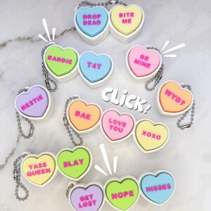 May include: A selection of heart-shaped keyrings in pastel colours, each bearing a different phrase in bold, bubble lettering. Phrases include "Drop Dead", "Bite Me", "Baddie", "T4T", "Bestie", "Be Mine", "WYD?", "Love You", "XOXO", "Yass Queen", "Slay", "Get Lost", "Nope", and "Misses".