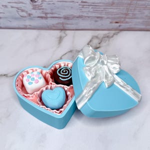 May include: A light blue heart-shaped box with a white ribbon bow, containing four chocolates. The chocolates include a white square with pink and blue dots, a dark chocolate swirl, and a blue heart-shaped chocolate.