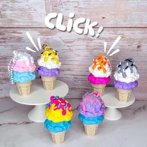 May include: A collection of colorful, decorative ice cream cone ornaments. Each cone features a waffle cone base, topped with layers of ice cream in various colors, and decorated with sprinkles and drizzles. The word "Click!" is written above the cones.