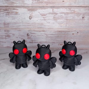 May include: Three black figurines with red eyes and small wings are displayed. Each figure has a loop at the top, suggesting they could be used as ornaments or keychains. The figures are standing on a white surface with a wooden background.
