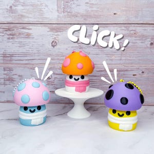 May include: Three colorful mushroom-shaped figurines with smiling faces. One is pink and blue, another orange and white with a pink scarf, and the third is purple and yellow. The word "CLICK!" is written above the figurines.
