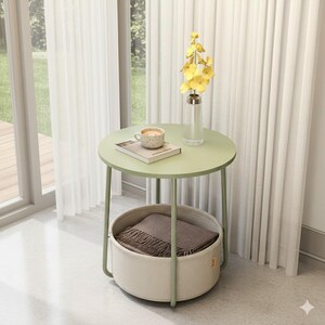 May include: A round, light green side table with a lower basket. The table has a light green top and legs, with a woven basket below. A book, a cup of coffee, and a vase of yellow flowers sit on top. A brown blanket is in the basket.
