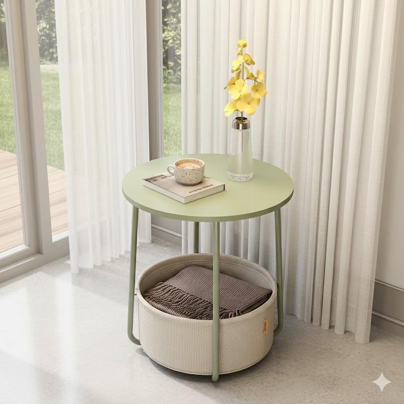 May include: A round, light green side table with a lower basket. The table has a light green top and legs, with a woven basket below. A book, a cup of coffee, and a vase of yellow flowers sit on top. A brown blanket is in the basket.