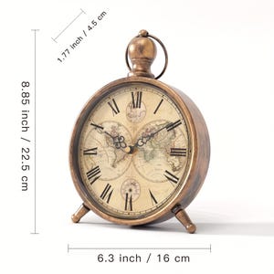 May include: A round, bronze-toned metal table clock with a vintage world map face, Roman numerals, and a top ring handle. It measures 22.5 cm in height, 16 cm in width, and 4.5 cm in depth, supported by two small front feet.