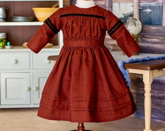 Puede incluir: Un vestido de manga larga en tela de color rojo óxido profundo con un pequeño patrón geométrico negro repetitivo. La prenda incluye un cuello alto, un corpiño ajustado con detalles de ribete negro en el pecho y las mangas, y una falda amplia.