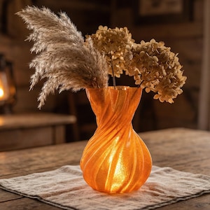 Könnte beinhalten: Eine orangefarbene Vase mit gedrehtem Design, die getrocknete Blumen und Pampasgras enthält. Die Vase steht auf einem neutralfarbenen Leinentuch auf einem Holztisch. Die warme Beleuchtung schafft eine gemütliche, einladende Atmosphäre.
