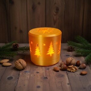 Könnte beinhalten: Ein zylindrischer, goldgelber Kerzenhalter mit ausgeschnittenen Weihnachtsbäumen und Sternen. Die Oberseite weist ein Schneeflockenmuster auf. Der Kerzenhalter wird von innen beleuchtet und wirft einen warmen Schein. Die Szene umfasst Tannennadeln, Nüsse und Tannenzapfen.