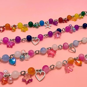 Op de afbeelding: Close-up van verschillende kleurrijke kralenarmbanden. Elke armband heeft een zilveren ketting met een verscheidenheid aan kralen in regenboogkleuren, waaronder roze, blauw, geel en groen. Sommige armbanden hebben ster- en hartbedels.