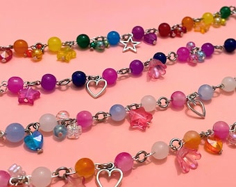 May include: Close-up of several colorful beaded bracelets. Each bracelet features a silver chain with a variety of beads in rainbow colors, including pink, blue, yellow, and green. Some bracelets have star and heart charms.