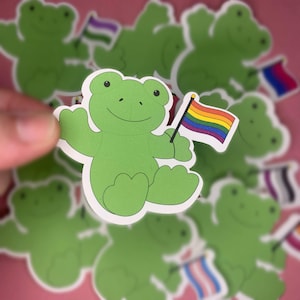 Peut inclure: Un autocollant découpé représentant une grenouille de dessin animé vert clair avec un visage souriant, tenant un petit drapeau à rayures horizontales rouges, orange, jaunes, vertes, bleues et violettes. L'autocollant a une bordure blanche.