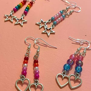 May include: A selection of beaded earrings with silver charms. One pair has rainbow beads and star charms. Another pair features pink and blue beads with star charms. The other two pairs have heart charms and pink, red, and purple beads.