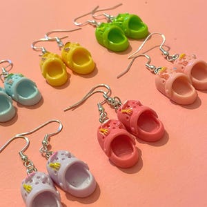 May include: A collection of colorful, miniature clog earrings in various shades, including green, yellow, pink, and blue. Each earring features a silver hook and is designed to resemble a tiny shoe. The earrings are displayed on a pink background.