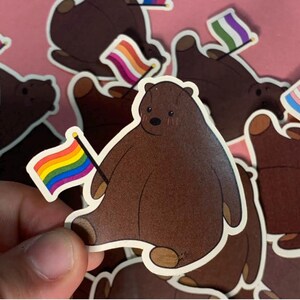 Pode incluir: Um autocolante recortado com um urso castanho de desenho animado com um corpo arredondado e orelhas pequenas, segurando uma pequena bandeira arco-íris num mastro preto. O urso tem uma cara simples com um nariz preto e olhos pequenos.