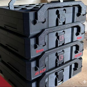 May include: Stack of black storage cases with handles and latches. Red text on the cases reads "9mm" and ".45 ACP". The cases are designed to hold ammunition and have a textured surface.