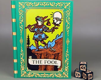 May include: A tarot card deck with a frog character labelled "THE FOOL." The card has a green border with gold filigree. Three dark blue dice with gold symbols are next to the deck. The card's illustration is on a yellow background.