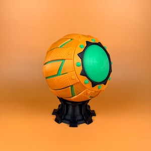 May include: A spherical, decorative object with an orange exterior featuring textured panels and green accents. The object has a large, circular green center and is mounted on a black base. The design incorporates geometric shapes and contrasting colors.