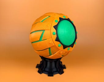 May include: A decorative sphere in orange with green and black details. The sphere has a textured surface and a large green central element. It is mounted on a black base. The design incorporates geometric shapes and contrasting colours.