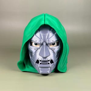 May include: A three-dimensional bust of a character wearing a metallic silver mask with visible rivets and a textured green hood. The mask features a rigid, angular design with a prominent nose piece, a segmented mouth area, and visible eyes.